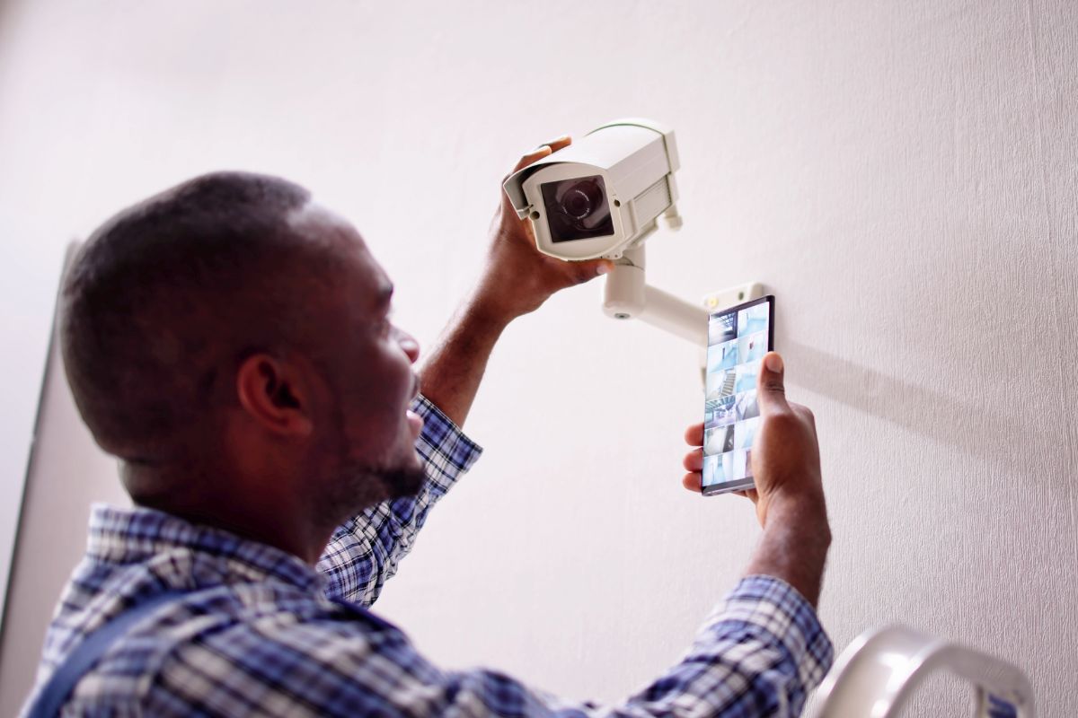 A person installs a security camera on the wall while holding a smartphone displaying surveillance footage.