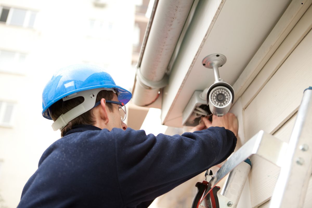 A worker in a blue hard hat installs a security camera on a building, using tools while standing on a ladder.