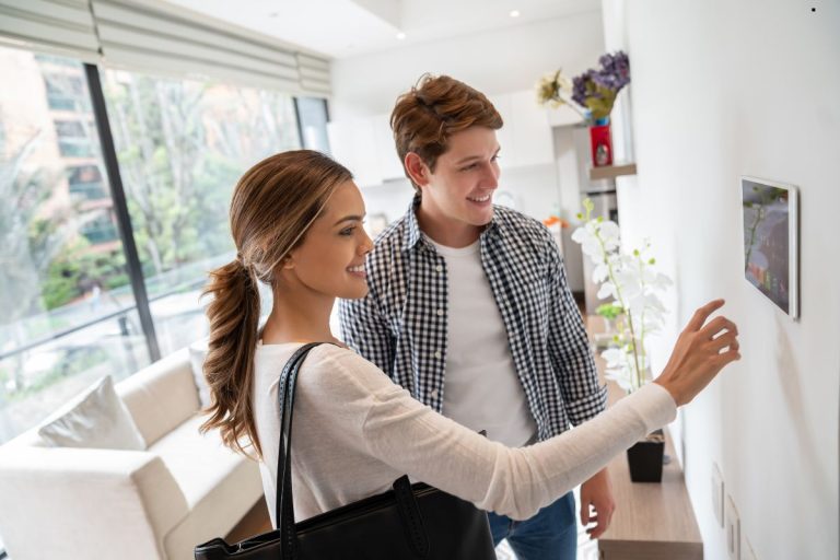 A woman in a white sweater adjusts a touchscreen in a modern living room while a man in a checkered shirt watches attentively.