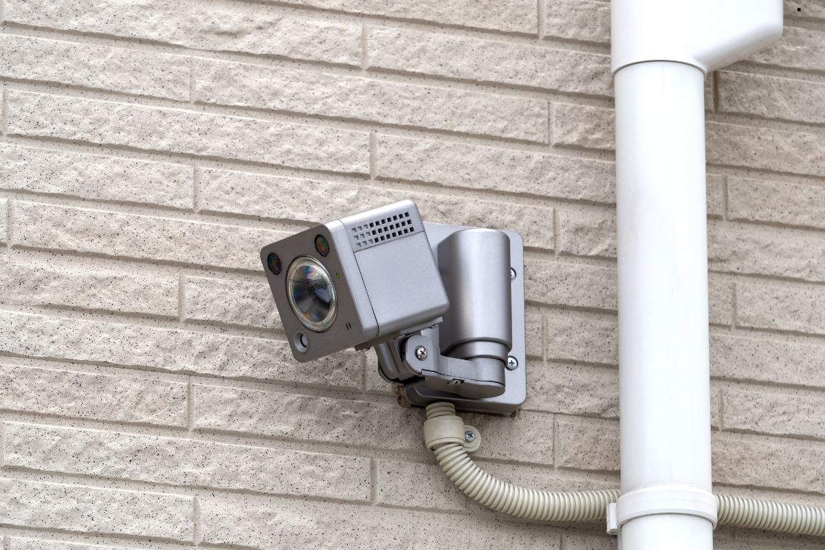 A mounted surveillance camera with a sleek silver design attached to a textured beige brick wall, connected by a light-colored cable.